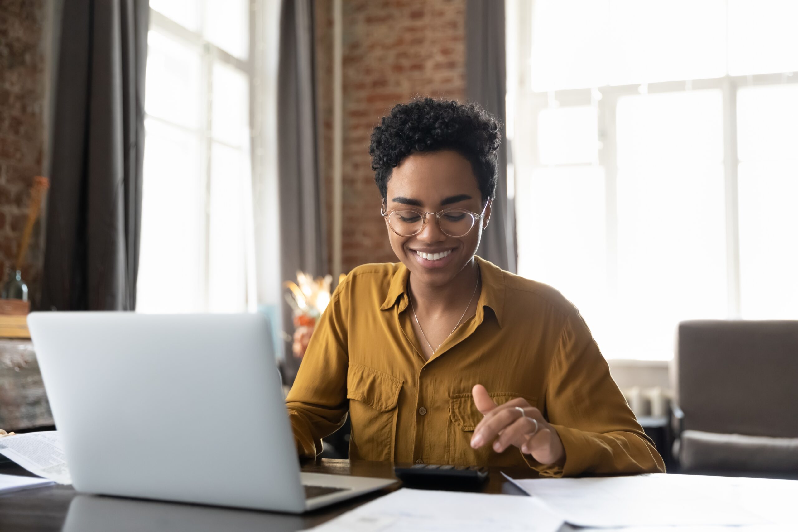 A young woman smiles as she types figures into a calculator, an open laptop on the desk in front of her.